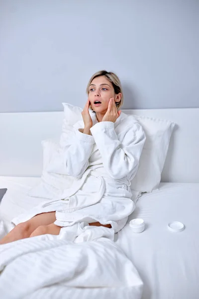 Mujer en albornoz aplicando crema en la cara tocando piel sensible hidratada y saludable — Foto de Stock