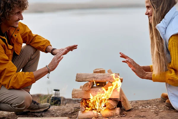 Young man and woman hands warm by fire. They sit around the fire. Travel concept. — Stock Photo, Image