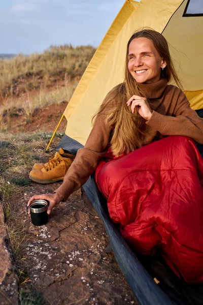 Donna in maglione bere il tè mentre seduto in tenda, in viaggio campeggio nella natura selvaggia — Foto Stock