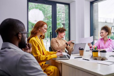 Group of diverse male and female colleagues gathering at table coworking, discussing business deal, at conference, dressed in stylish formal clothes. cups of coffee on table, creative team
