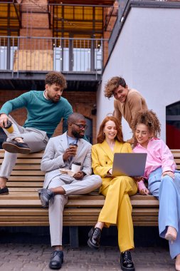 business people relaxing, have conversation and sitting on bench outdoors, drinking coffee, surfing the Net. caucasian and african coworkers in trendy elegant clothes enjoy spending time outdoors