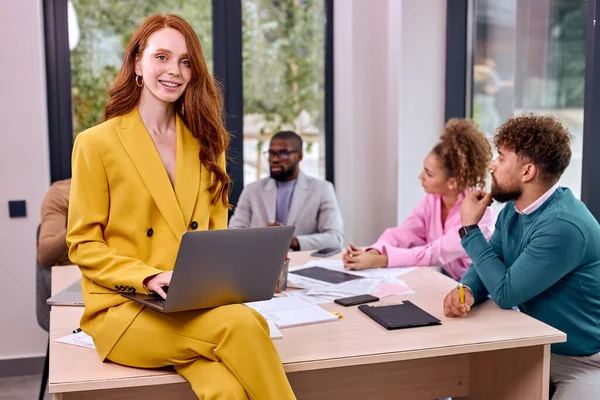 Portrait of redhead excited woman in yellow formal wear sitting on desk with laptop with colleagues in the background, excited caucasian female enjoy working in office with, creative coworking