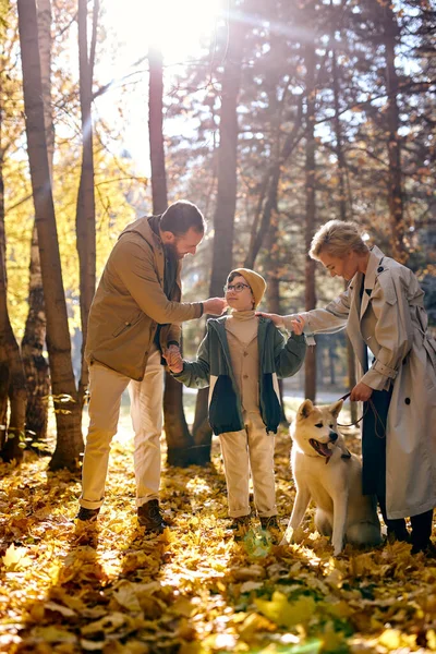 caucasian european family in warm clothes walking with son with white pet in autumn forest or park at sunny day, man woman and child happy together. Family, friendship, caring, healthy communication.