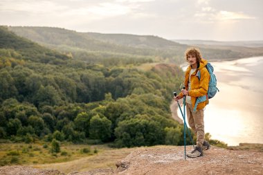 active man trekking on mountains having relaxing break, holding hiking poles, in contemplation of landscape nature surrounding him. curly caucasian male in sportive outfit enjoy hiking
