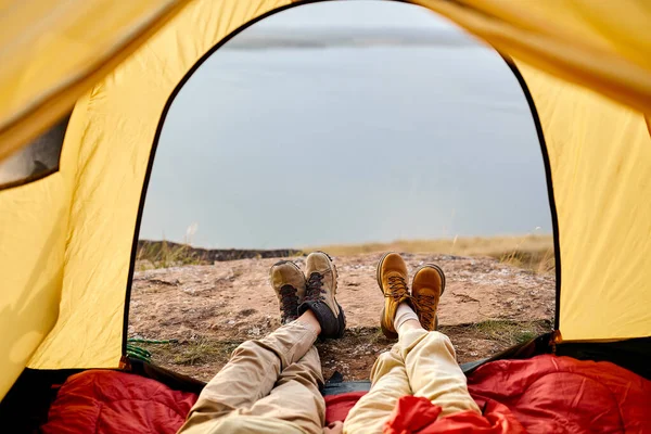 High angle view of cropped couple camping at lake on autumn day, lying at tent entrance, cuddling and enjoying beautiful sunset over the lake, relaxed man and woman in casual wear have rest