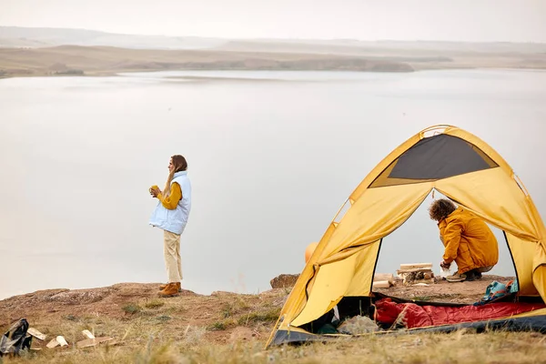 couple travelling in wild nature by lake river. man preparing tent and bonfire closer to the night. caucasian man and woman in casual wear in nature, woman drinking tea and enjoying the landscape