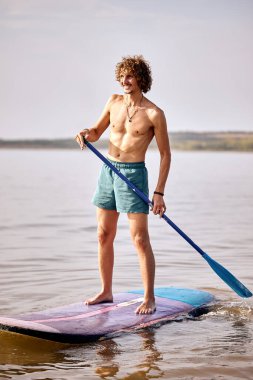 shirtless young caucasian male standing on surfboard in river, concentrated. man during active surfing training alone. Surfing, subsurfing, active sport concept