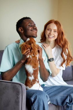 Joyful black man and caucasian woman sit on sofa embarrassing playing with pet, redhead lady and black american guy in casual outfit have rest, enjoy weekends, spend time together indoors