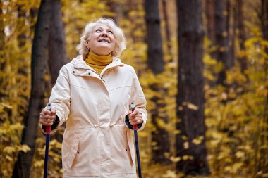 European aged lady with gray short hair standing in forest with Nordic walking sticks, going home after training, smiling broadly and looking at side, in contemplation. recreation in park.