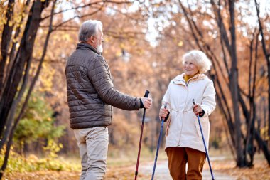 Senior aged couple walking with nordic walking poles in autumn park. Mature woman and old man doing exercise outdoors. Healthy lifestyle concept. Caucasian retired male and female enjoy the walk