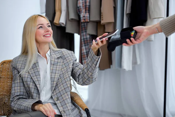 Mobile Shopping. Young Woman Using Phone Paying For New Dress, Fashion Trends Outfits In Background. Fashion And Style Application For Smartphone. Blonde Lady In Gray Jacket Smiling Looking At Seller
