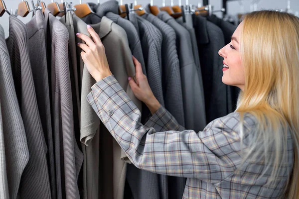 Female looking at new collection of stylish clothes on hangers clothing rack rails, fashion design studio store concept. blonde caucasian female in jacket make choice, young customer woman