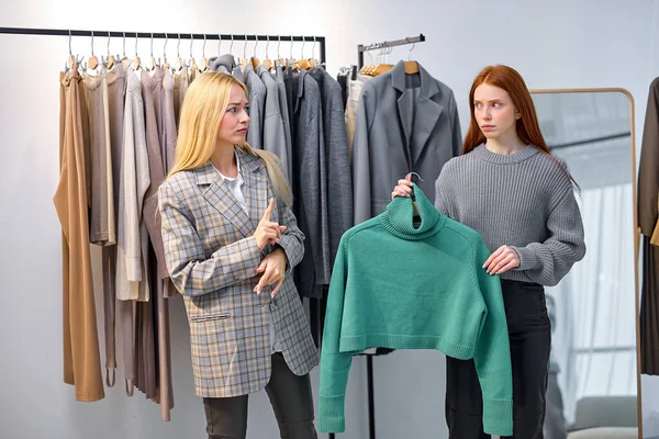 Young caucasian fashion consultant and dubious lady having talk discussing new trends in world of fashion. redhead woman holding green shirt on hanger, make choice. in cozy modern showroom
