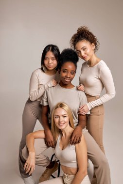 Positive smiling women with diverse appearance and natural make-up posing together, attractive ladies in casual clothes look at camera having cute smile on face, isolated on beige background