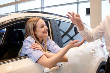 Beautiful young caucasian woman getting key and smiling while sitting in new car in dealership, cropped male manager in formal wear giving keys by white car automobile, woman make purchase