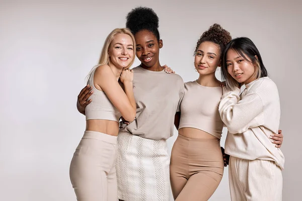 beauty portrait of diverse women, caucasian, asian and african american ladies with different skintone posing at camera, hugging each other, dressed casually in beige tone clothes. isolated