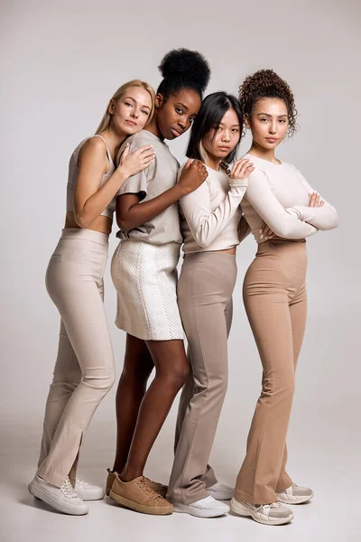 Female Beauty. Four Friendly Women Looking At Camera Posing In Studio On White Background. Isolated. Indoors. Gorgeous Calm Pacified Ladies In Casual Modern Outfit Together, Hugging