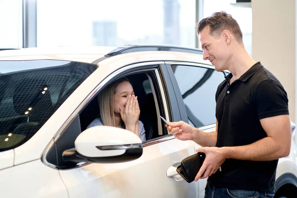 handsome guy buying a new car for caucasian cheerful female at dealership, pleasant excited lady sit inside of car shine with happiness. in cars showroom. consumerism, auto, transport