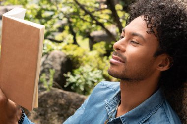 Afro-Amerikan adam kitap okuyor ve parkta doğanın tadını çıkarıyor.