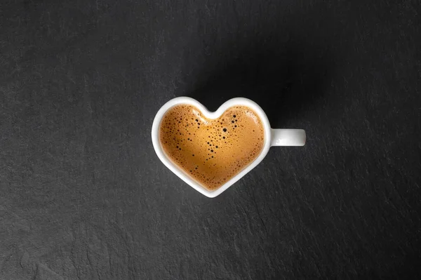 White cup in the shape of a heart with coffee and beautiful foam on a black stone background. view from above. copy space — Stock Photo, Image