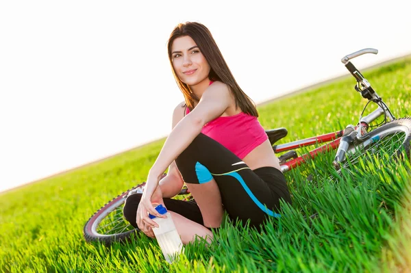 Mujer joven descansando con bicicleta en el hermoso campo sosteniendo una botella de agua — Foto de Stock