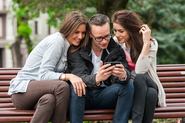 Three friends looking at an image on a smartphone while sitting on a park bench — Stock Photo, Image