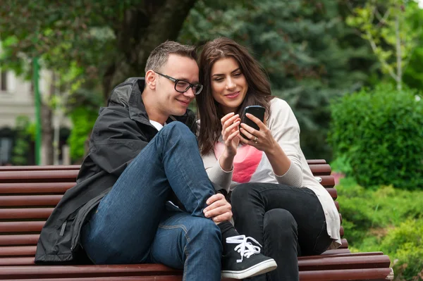 Dois amigos compartilhando um smartphone em um banco de parque — Fotografia de Stock