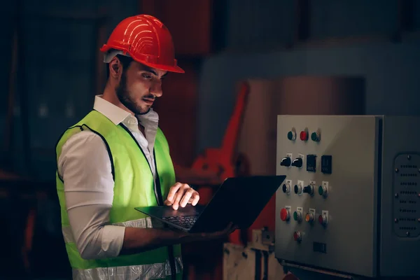 Fabrika işçisi, endüstriyel şantiyede fazla mesai yapan dizüstü bilgisayarla elektrik ve makine işlemlerini kontrol ediyor.