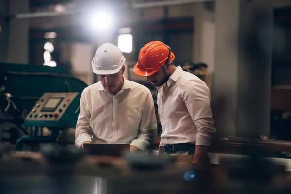 Fabrika işçisi ve mühendisi metal levha endüstriyel şantiyesinde birlikte çalışıyorlar, bilgisayardaki makine işlemini kontrol ediyorlar ve güvenlik için başlık takıyorlar.