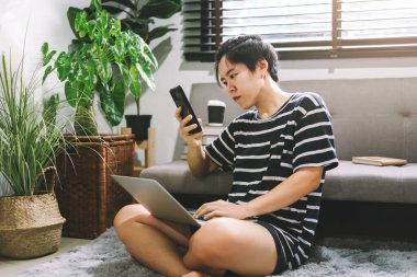 Lezbiyen LGBTQ Asyalı kadın evden iş için akıllı telefon ve laptop kullanıyor. Oturma odasında covid-19 durumu, günlük takım elbise giyiyor..