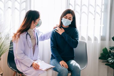 Asyalı doktor muayenehanedeki genç yetişkin kadını muayene ediyor. Doktor, kovid-19 salgınını korumak için maske takıyor ve sağlık hizmetlerini kontrol ediyor.