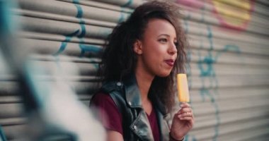 Afro-Amerikan kız bir vintage Bisiklet şehir kaldırım gülümseyen üzerinde oturan ve uzak bir portakal aromalı Buz lolly, ağır çekim yaparken seyir ön planda geçmiş gördüm gülümseyerek