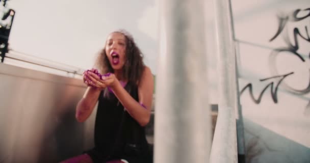 スローモーションでカメラに向かって彼女の手のひらから明るいピンクのグリッターを吹く屋外階段に座っているアフリカ系アメリカ人の十代の少女 — ストック動画
