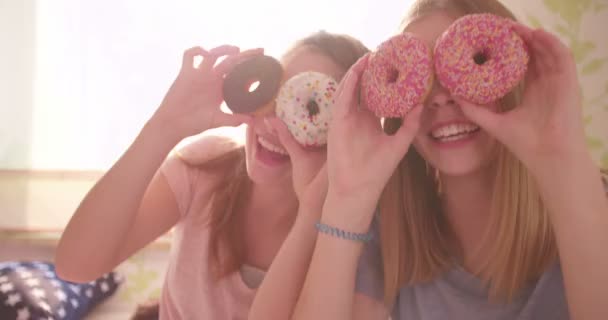 两个青少年女孩朋友拥有一套睡衣方在阳光明媚的卧室里 抱着色彩缤纷的甜甜圈到他们的脸 伸出舌头在相机同时遮住自己的眼睛 — 图库视频影像