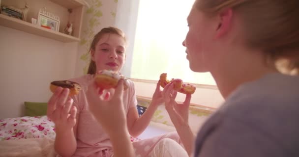 十代の少女は彼女の友人がドーナツを食べても 太陽に照らされた寝室で昼間のパジャマ パーティーの最中に彼女の摂食は甘いドーナツからかみ傷を取るを笑ってください — ストック動画