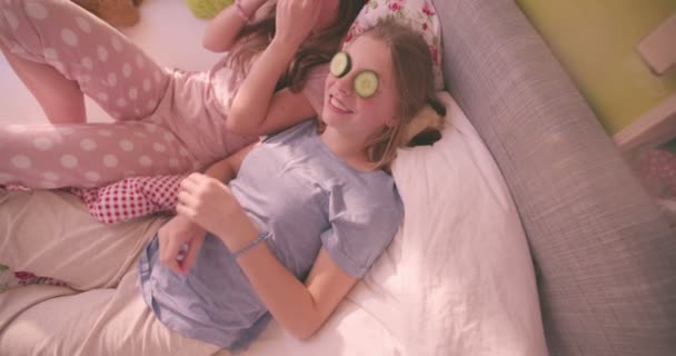 彼らが笑っている間 彼らの目に休んでいるキュウリの新鮮なスライスの快適パジャマでベッドに横たわっている 人の十代女の子のハイアングル ショット — ストック動画