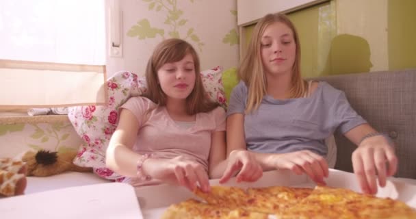 代の女の子を持つ パジャマ パーティー笑みを浮かべて 彼らの前にベッドの上のピザの大きいの持ち帰り箱と一緒にベッドに座りながら自分の顔の前で安っぽいピザの巨大なスライスを押し — ストック動画