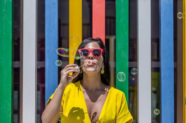Güneşli bir günde Latin bir kadının sarı bir elbise ve güneş gözlüğüyle sabun üfleyişini ön planda tutmak..