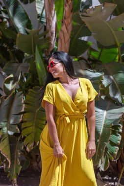 Dikey görüntüde, sarı elbiseli, güneş gözlüklü, ayakta duran ve yeşil çiçekli bir arka plana bakan Latin bir kadın var. Güneşli bir gün..