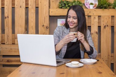 Resmi kıyafetli, gülümseyen, serbest çalışan, sıcak kahve içen ve kafede seramik tabakta tatlı makaronla netbook üzerinde çalışan genç bir kadın.