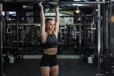 Spor tişörtlü, formda ve şortlu genç ve güçlü bir bayan atlet. Kafasında ağır bir plaka var. Modern spor salonunda basın egzersizi yaparken başka tarafa bakıyor.
