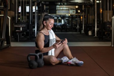 Spor kıyafetleri içinde uzun saçlı, omuzlarında havluyla gülümseyen, spor sonrası kettlebell 'in yanında oturan akıllı telefonuyla mesajlaşan genç, kaslı sporcu bir vücut.