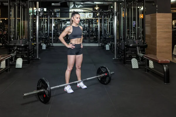 Spor kıyafetleri içinde uzun at kuyruklu, motive olmuş genç bayan sporcu elleri belinde ve spor salonunda ağır halteri kaldırmadan önce başka tarafa bakıyor.