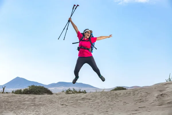 Positive female explorer with backpack and trekking poles jumping above sandy Famara beach and celebrating achievement