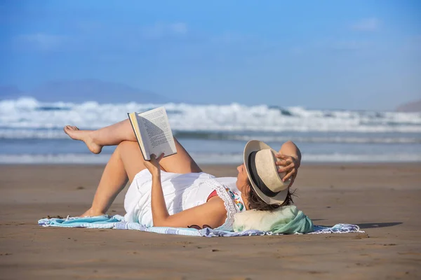 夏天的周末 在度假胜地 女游客在海浪和蓝天的映衬下 一边在沙滩上触摸帽子一边看书 — 图库照片