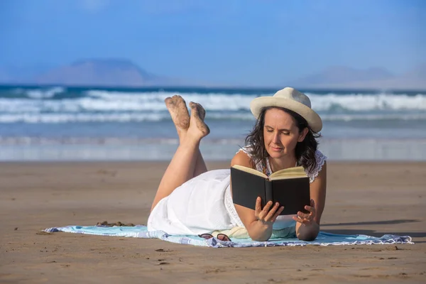 在夏天的周末 穿着白衣 头戴白帽的中年女性游客在沙滩上悠闲自在地散步 在海滨和蓝天的映衬下 阅读一本有趣的书 — 图库照片