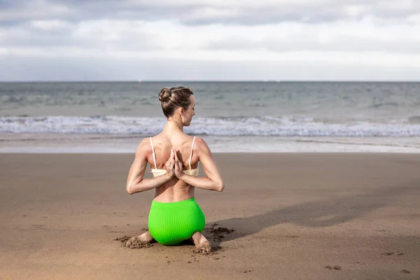 Slim female in bra and skirt clasping hands behind back while kneeling on sand near stormy sea during yoga session on beach