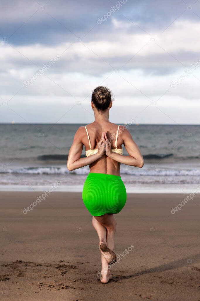 Anonymous female in skirt and bra clasping hands behind back while doing Garudasana pose on sandy beach on stormy day