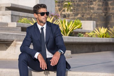 Serious male executive in suit and sunglasses sitting on concrete step and looking away on sunny day on city street