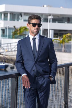 Confident businessman in suit and sunglasses holding hand in pocket and looking away while leaning on bridge railing on sunny day in city port.